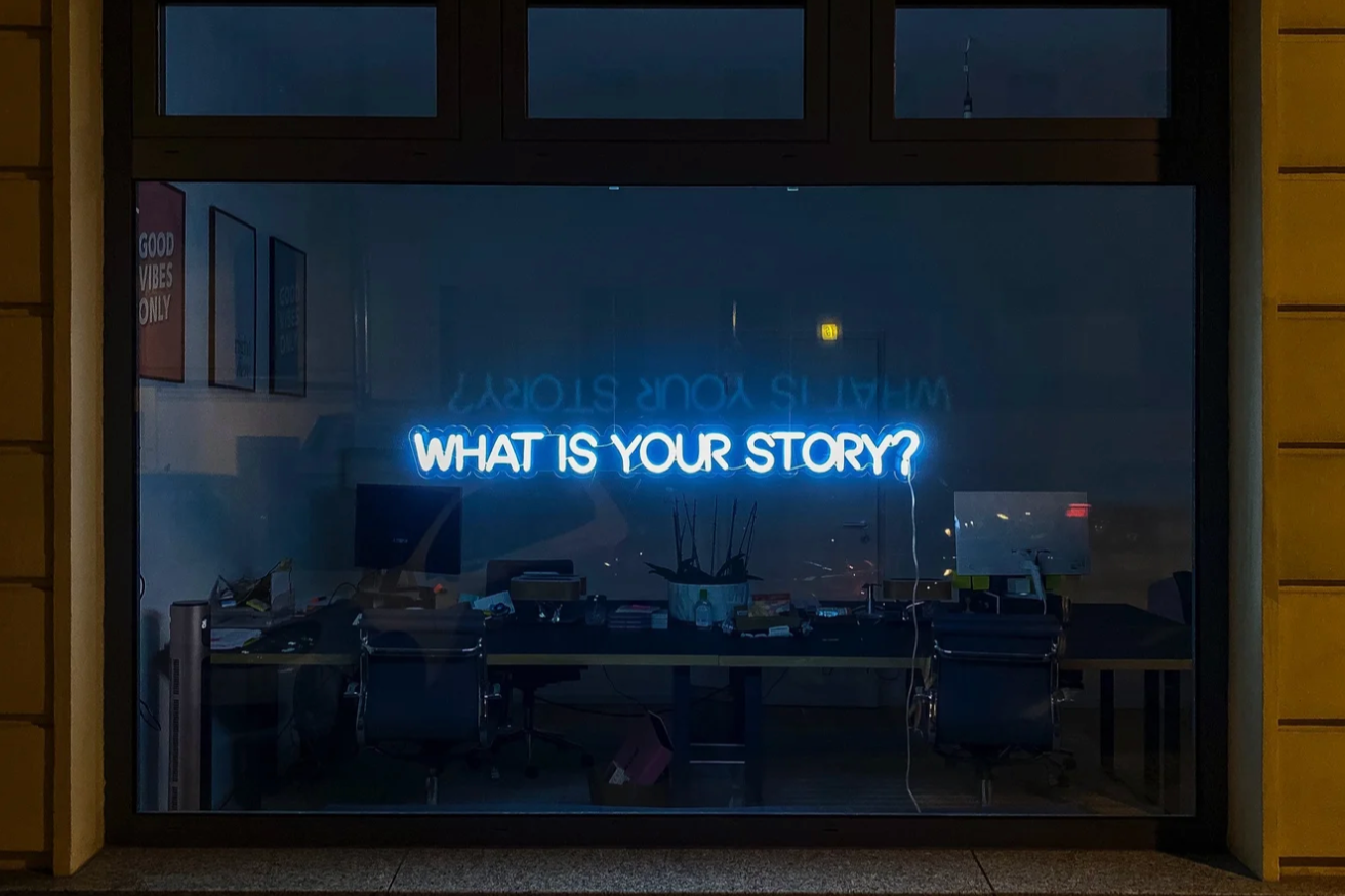 Neon sign in a window displaying the question, "What is your story?" with an office desk, chairs, and framed artwork visible inside.