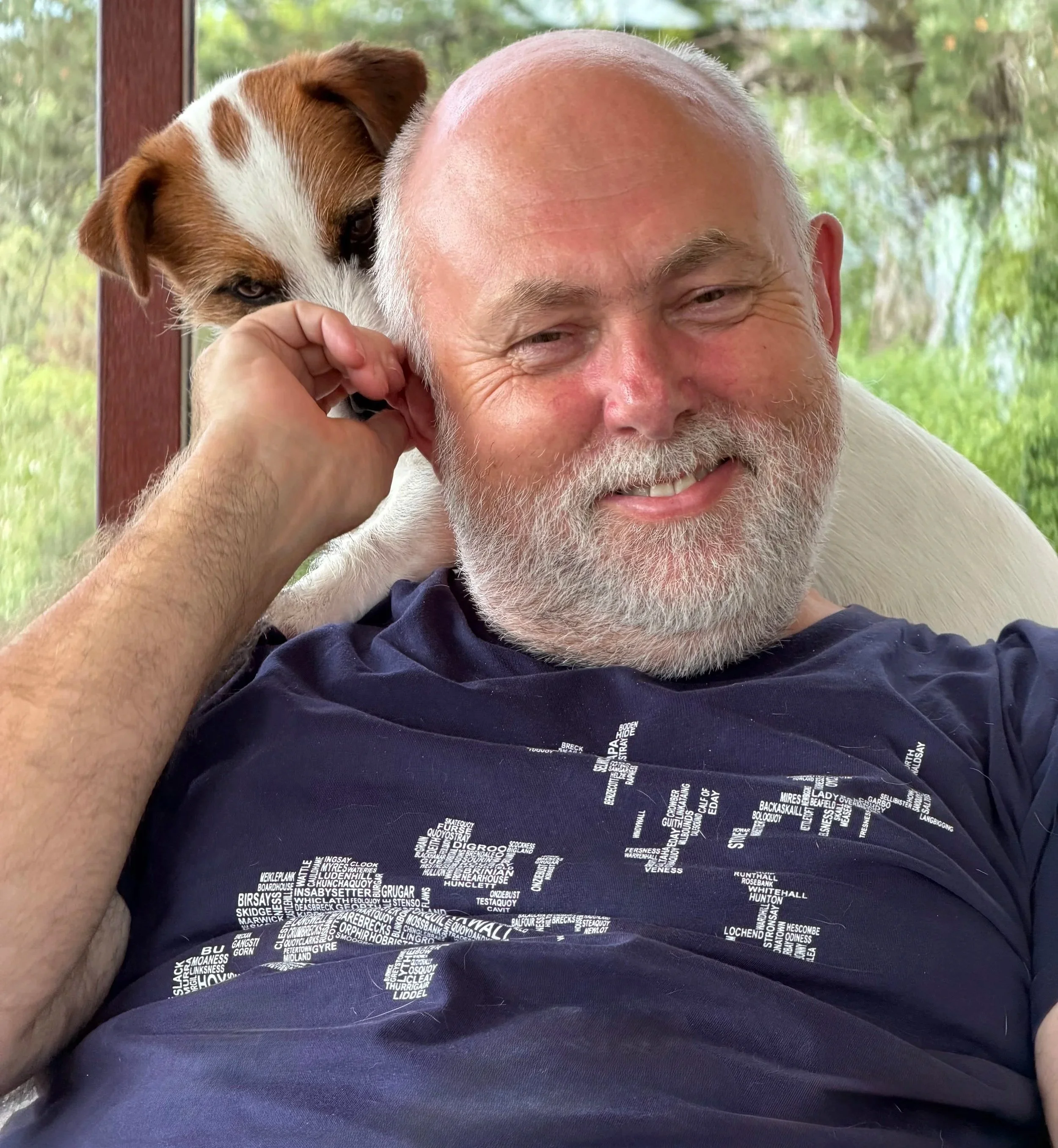 A man with a beard smiling while holding a dog close to his face. The dog is looking at the camera. They are indoors near a window with trees visible outside.