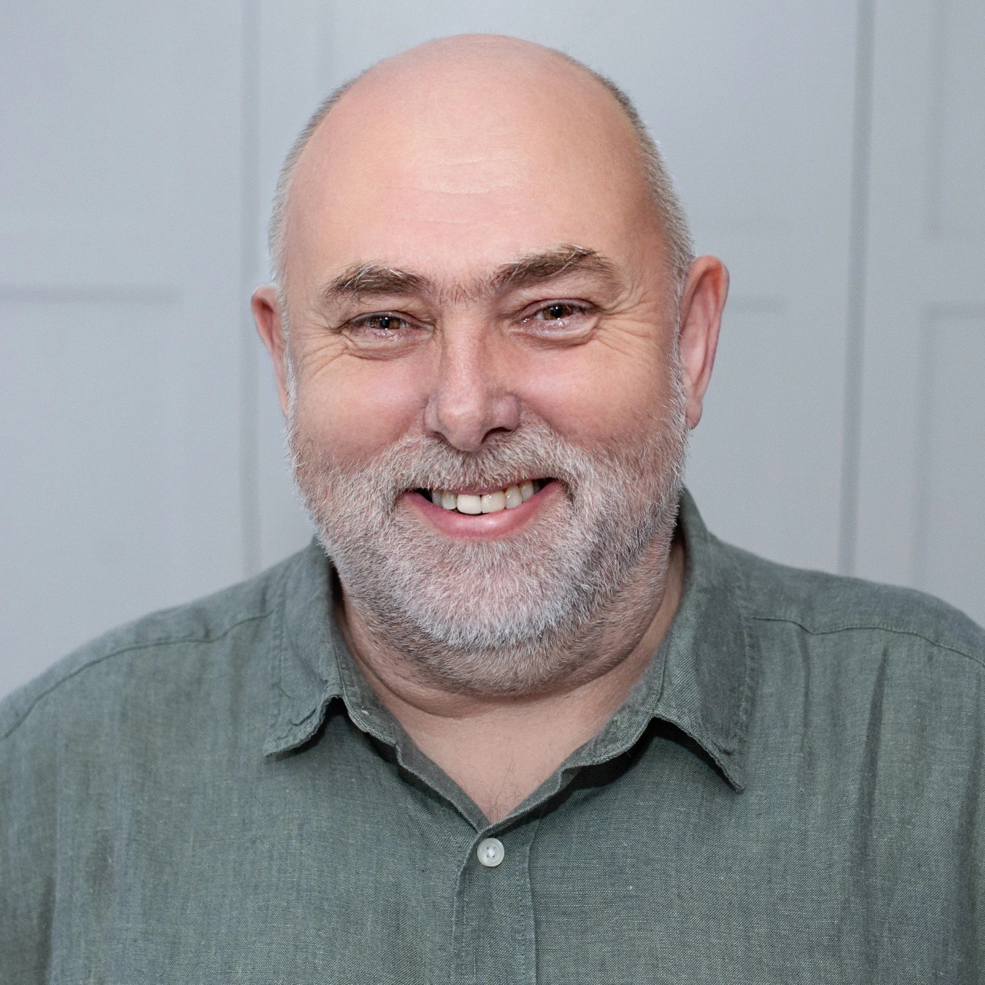 Close-up of a smiling middle-aged man with a gray beard, wearing a dark gray collared shirt, and standing against a plain light gray background.