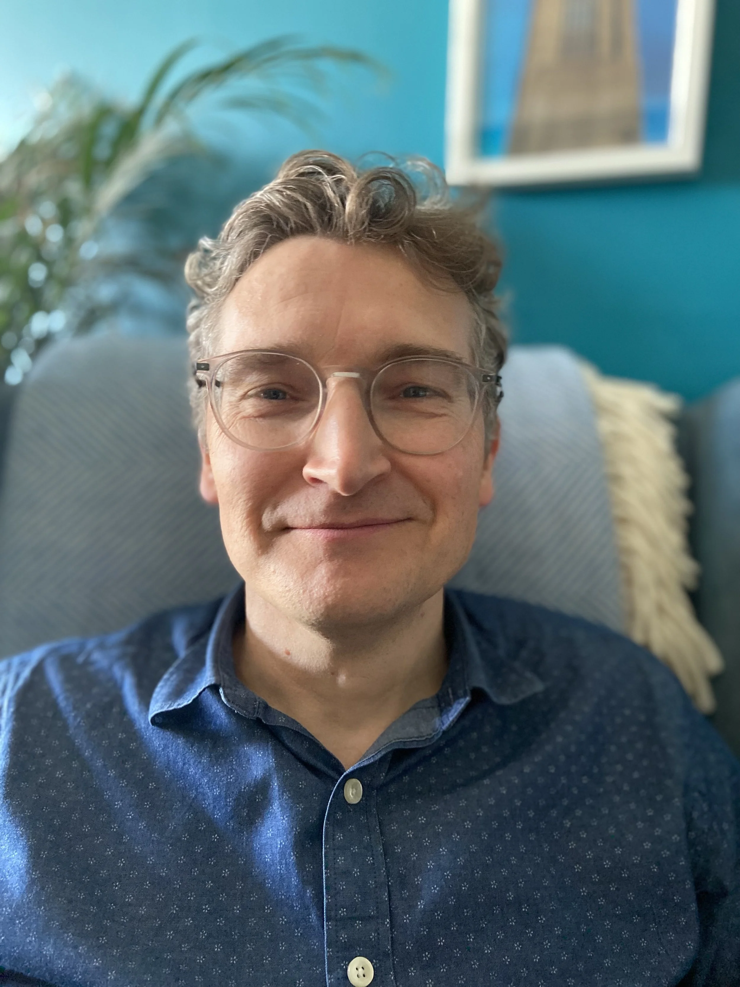A man with short, curly, gray hair and glasses smiling, wearing a dark blue patterned shirt, sitting on a couch with a light-colored pillow, in front of a teal wall with a framed picture and a potted plant.