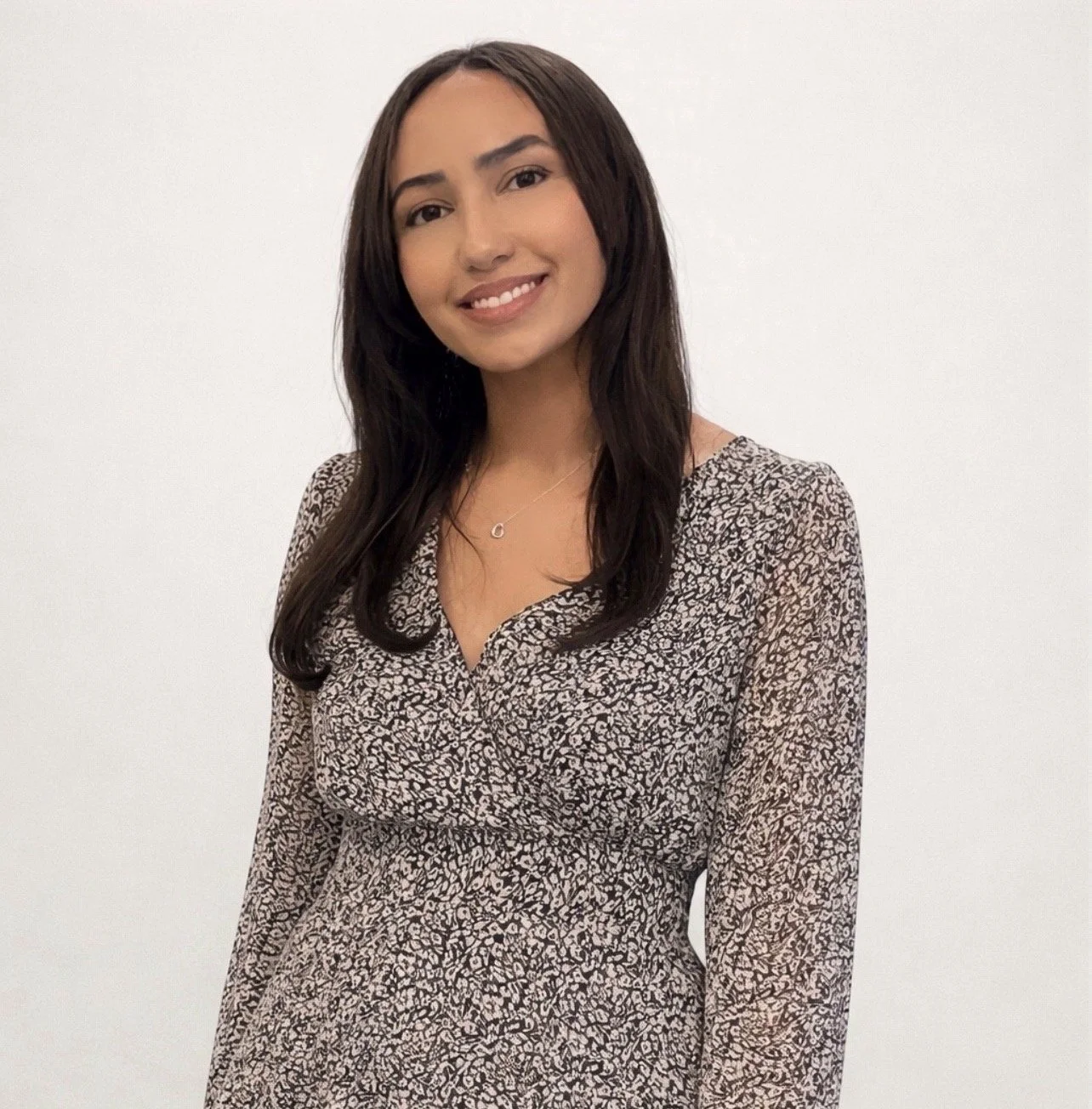 A young woman with long dark hair smiling, wearing a patterned dress and a delicate necklace, standing against a plain white background.