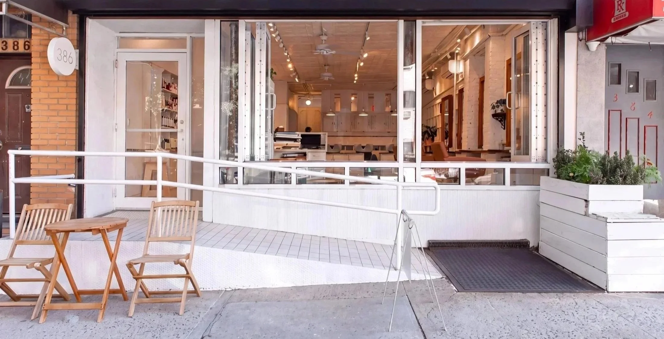 Modern restaurant storefront with open glass doors, outdoor wooden café chairs and table, wheelchair ramp, and planter boxes