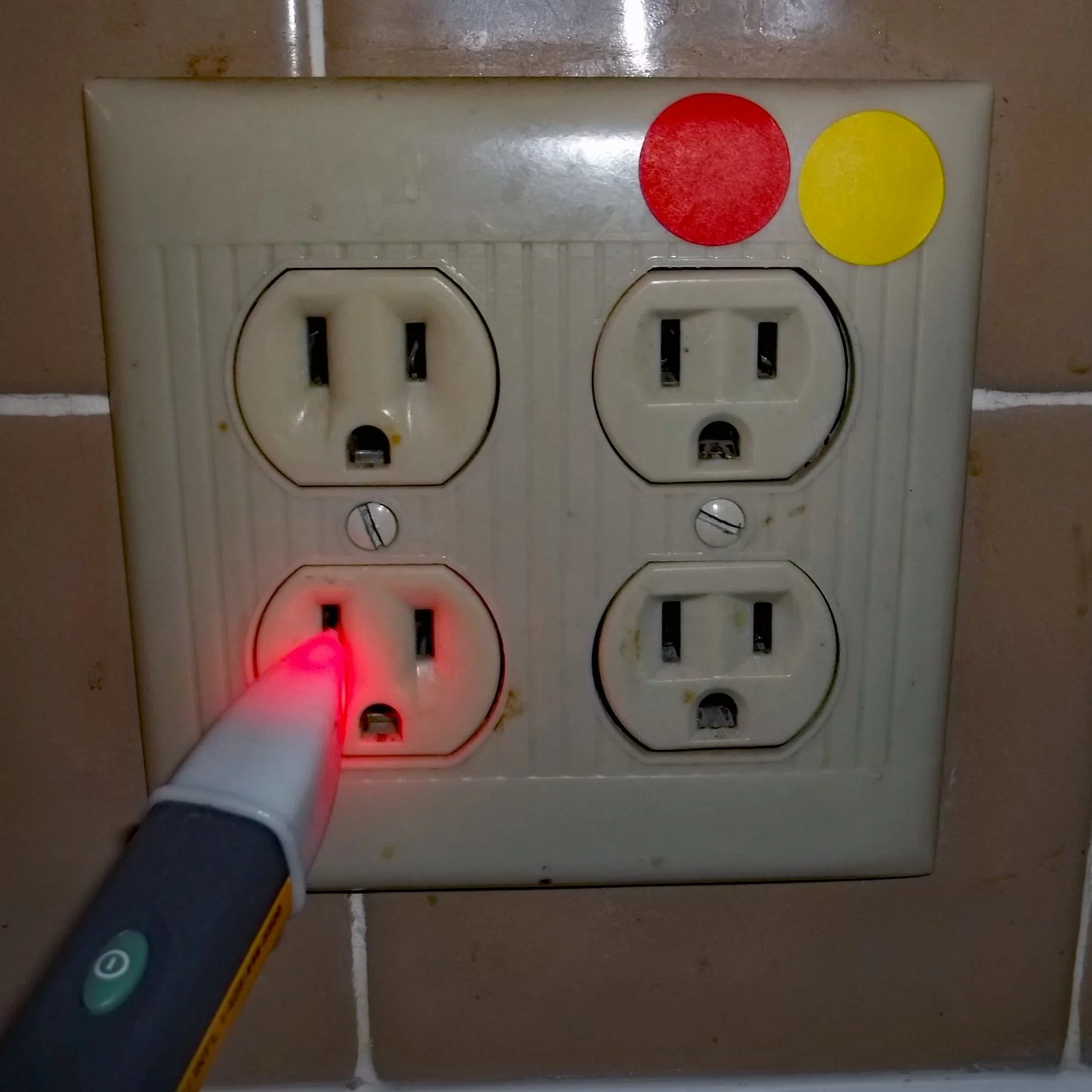 A person's foot in a sock is plugged into a white electrical socket on a tiled wall. The socket has three outlets, with the bottom left outlet currently in use by an electrical device emitting a red light. Two circular stickers, one red and one yellow, are adhered to the panel above the outlets.