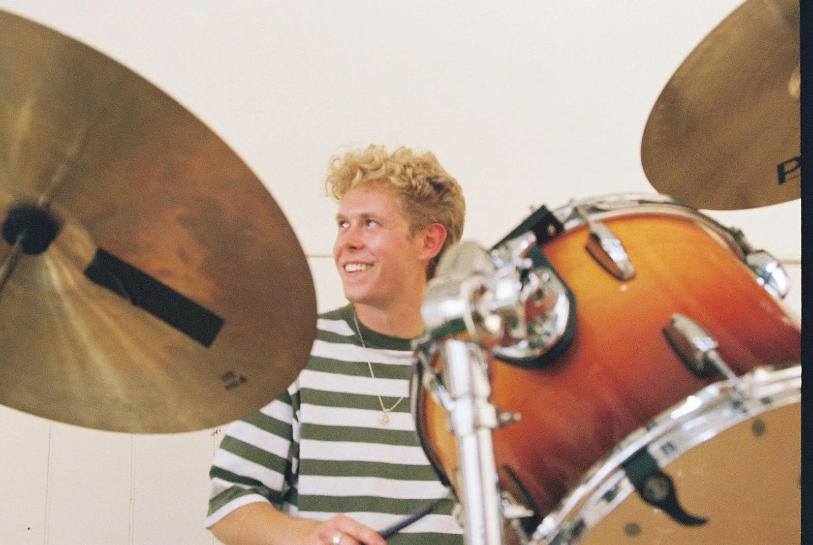 Young man with curly blonde hair smiling while playing a drum set, wearing a black and white striped shirt.