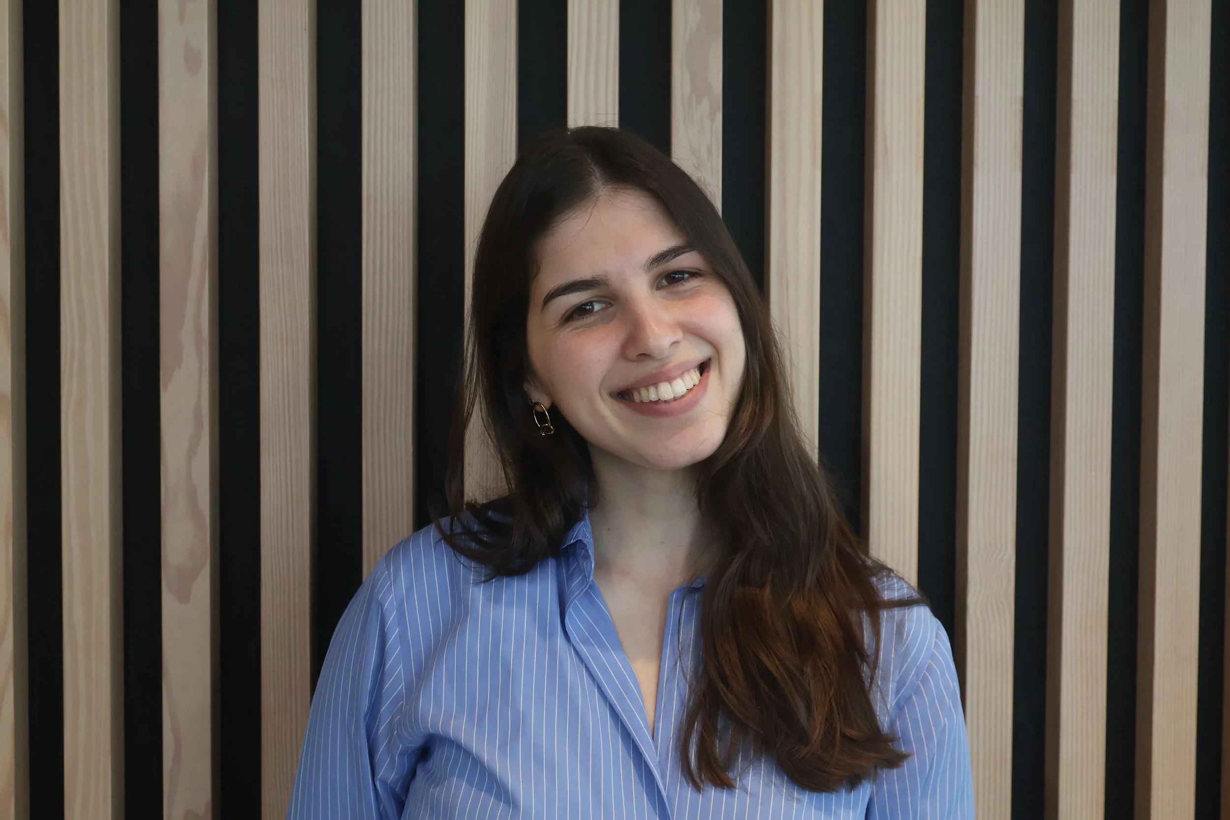 Mulher sorridente com cabelo castanho, usando camisa azul de listras brancas, em frente a uma parede de painéis de madeira.