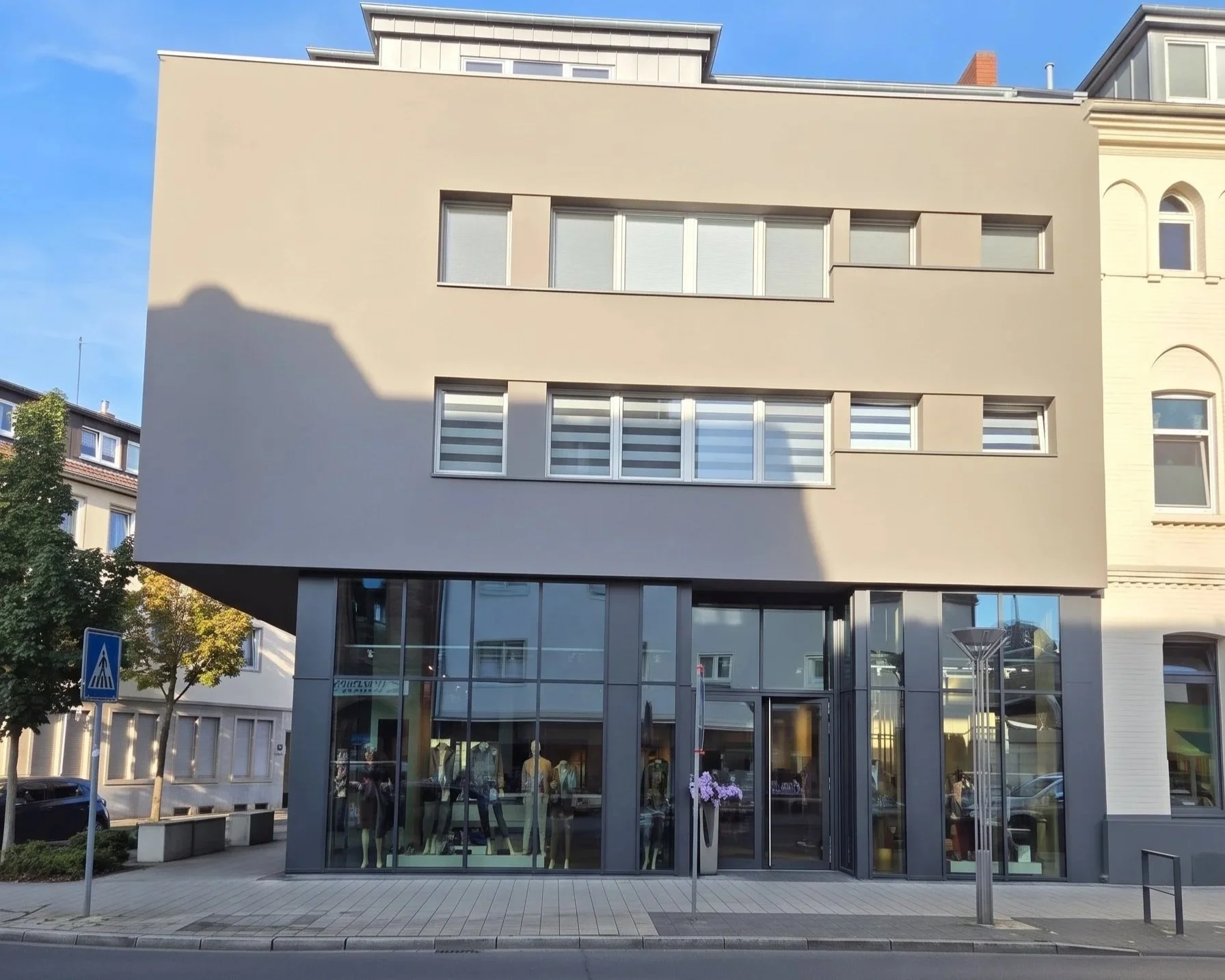 Moderne Einkaufsgeschäft mit großen Glasfenstern im Erdgeschoss, Bäumen und Fußgängerüberweg vor dem Gebäude, eher städtische Umgebung bei Tageslicht