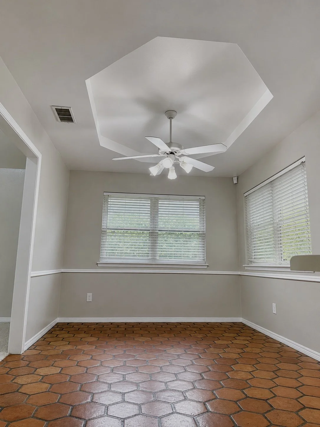 Empty room with beige walls, white trim, hardwood floor in a hexagonal pattern, double windows with blinds, and a ceiling fan with lights.