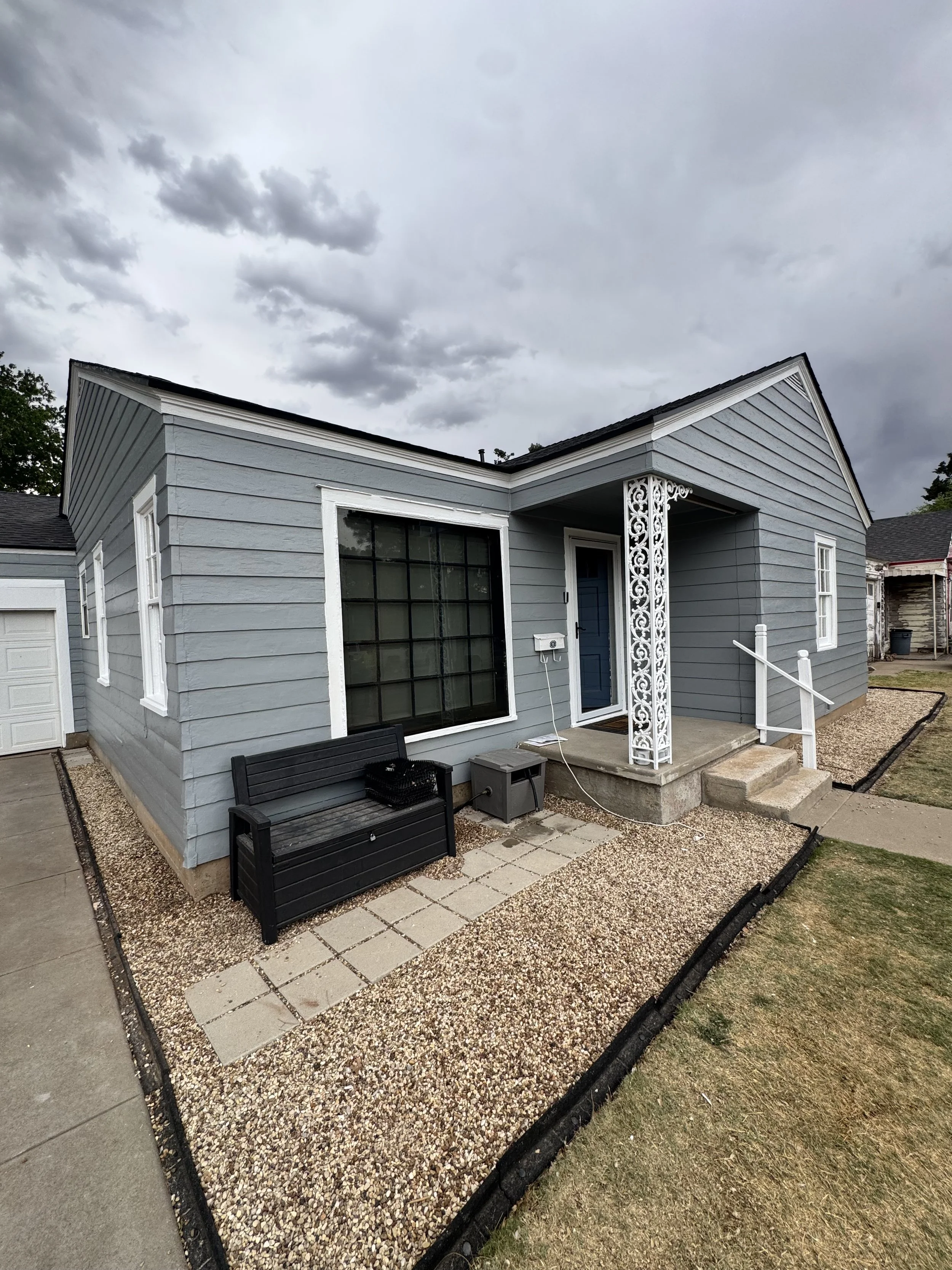 A house with blue siding and white trim, a small porch with steps, a black bench, and a gravel and paver patio, under a cloudy sky.