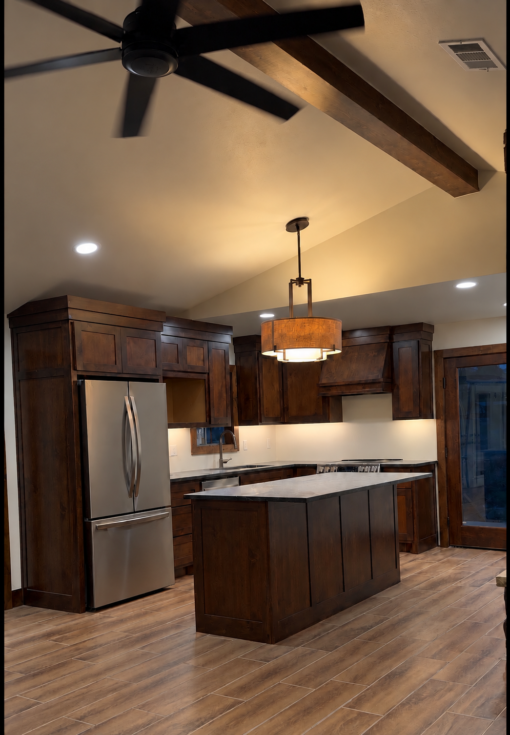 Kitchen with dark wood cabinets, stainless steel refrigerator, marble countertop island, and hardwood floors. Includes a ceiling fan, a pendant light, and a sliding glass door.
