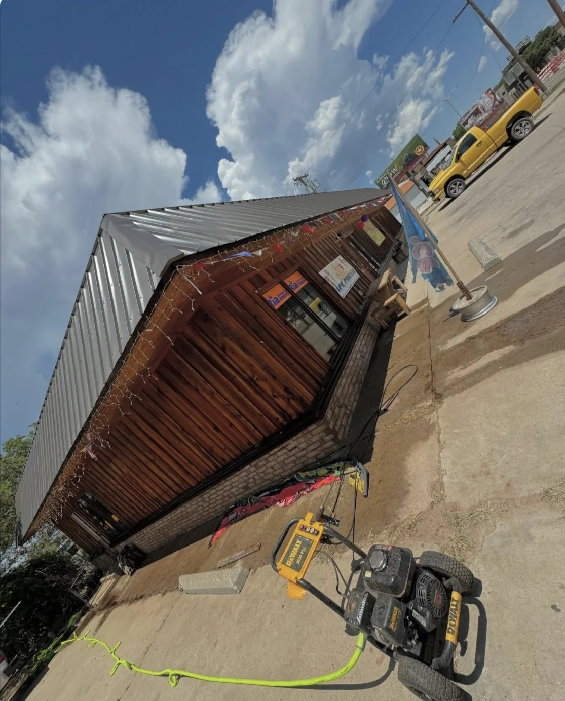 A small wooden building with a metal roof decorated with string lights and an ice flag outside. A pressure washer is on the ground, and a yellow pickup truck is parked nearby on a sunny day with partly cloudy skies.
