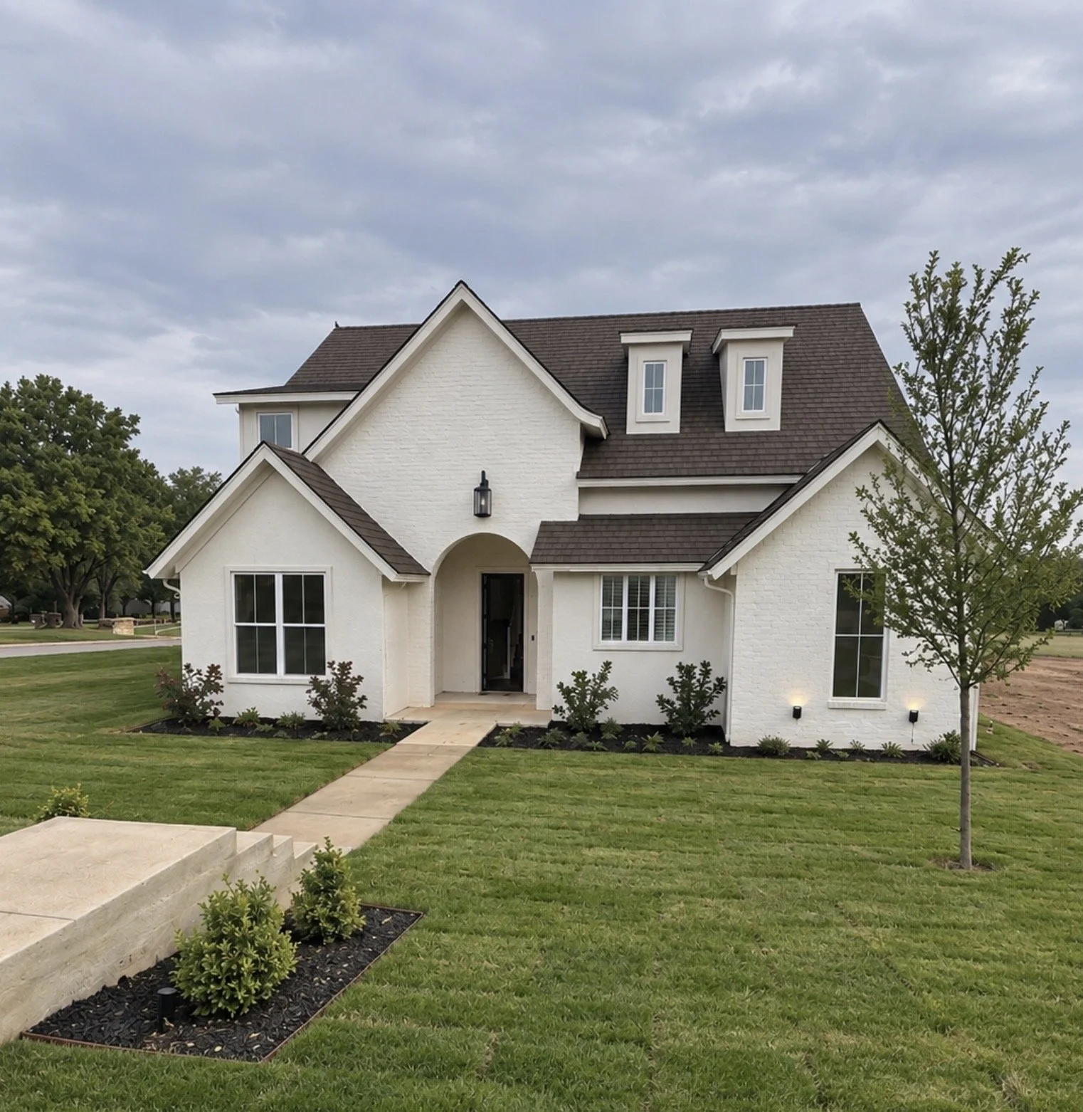 Front view of a white two-story house with dark roofing, large windows, a front porch with an arched entrance, and landscaped lawn with small plants, a sidewalk, and a young tree on the right.
