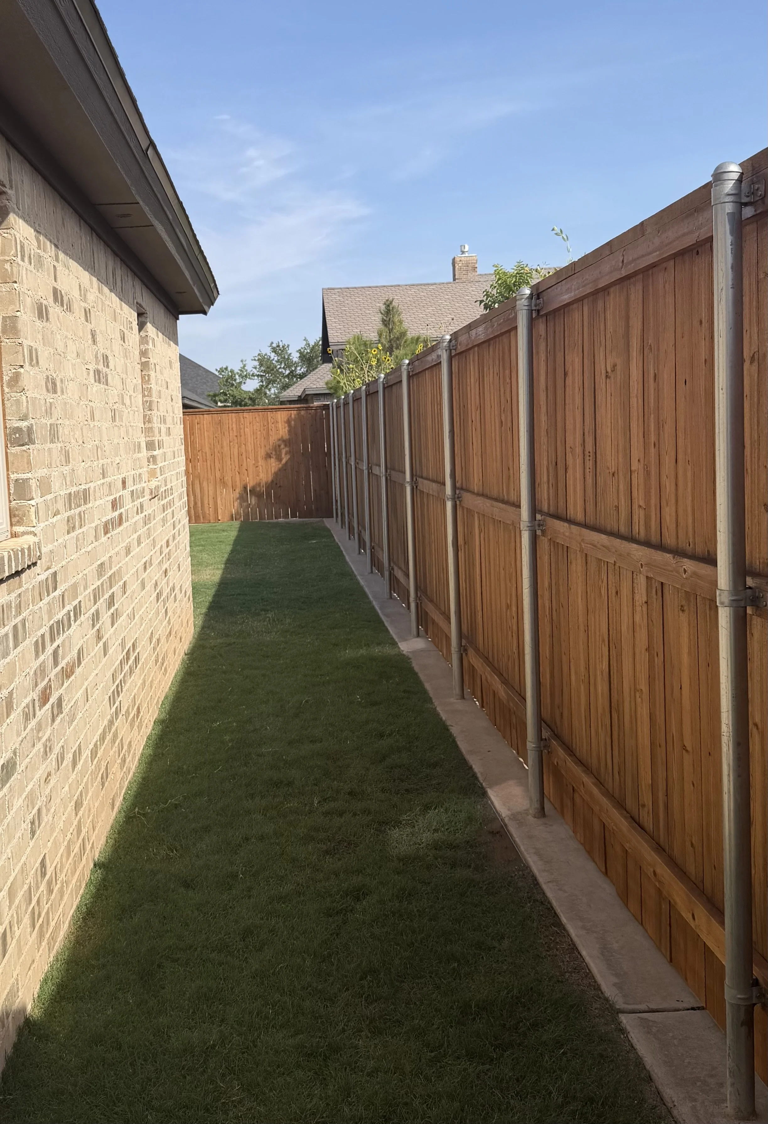 Side yard with a brick house on the left and a wooden fence on the right, green grass in between, and a clear blue sky above.