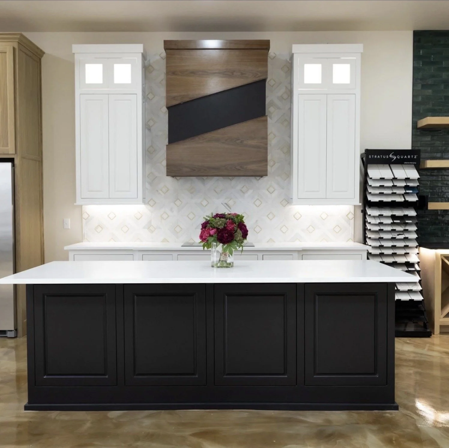 Modern kitchen island with a black base and white countertop, topped with a vase of pink and purple flowers, set against white cabinet wall with decorative tiles, flanked by white wall cabinets and a display of tile samples on the right.