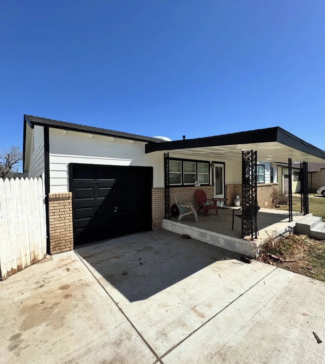 Front view of a house with a black garage door, a covered porch with outdoor furniture, and a concrete driveway under a clear blue sky.