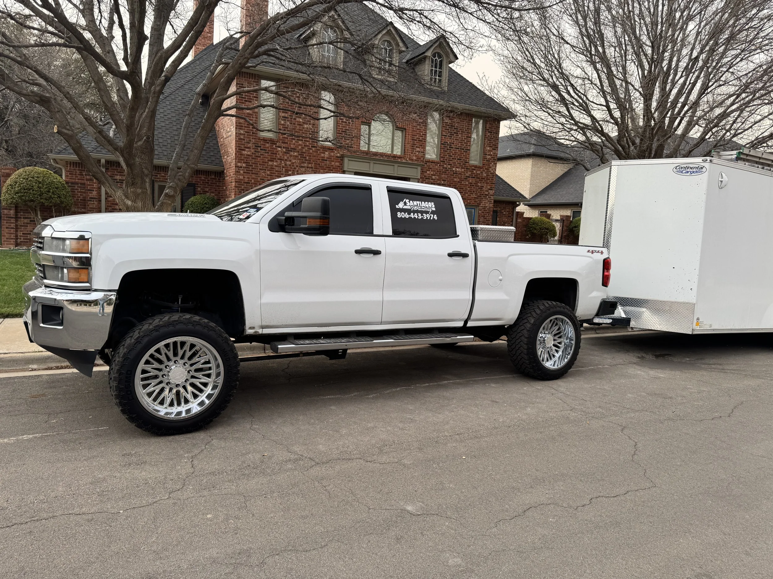 A white pickup truck with large chrome wheels parked on a residential street in front of a brick house with trees.