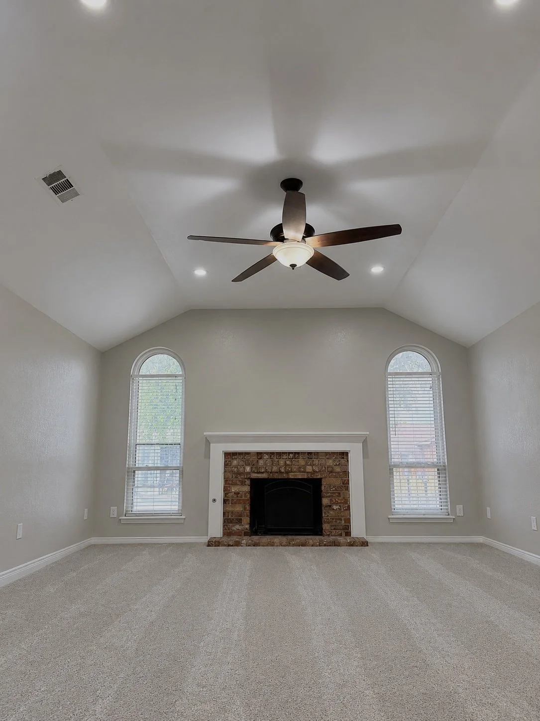 Empty living room with beige carpet, stone fireplace, two tall arched windows, ceiling fan, and recessed lighting.