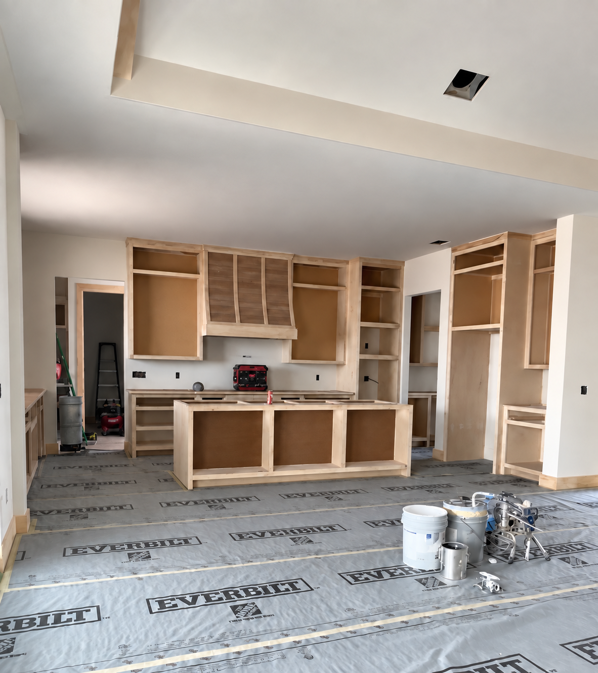 Kitchen under construction with wooden cabinets and shelves being installed, construction tools and materials on the floor, and unfinished walls.