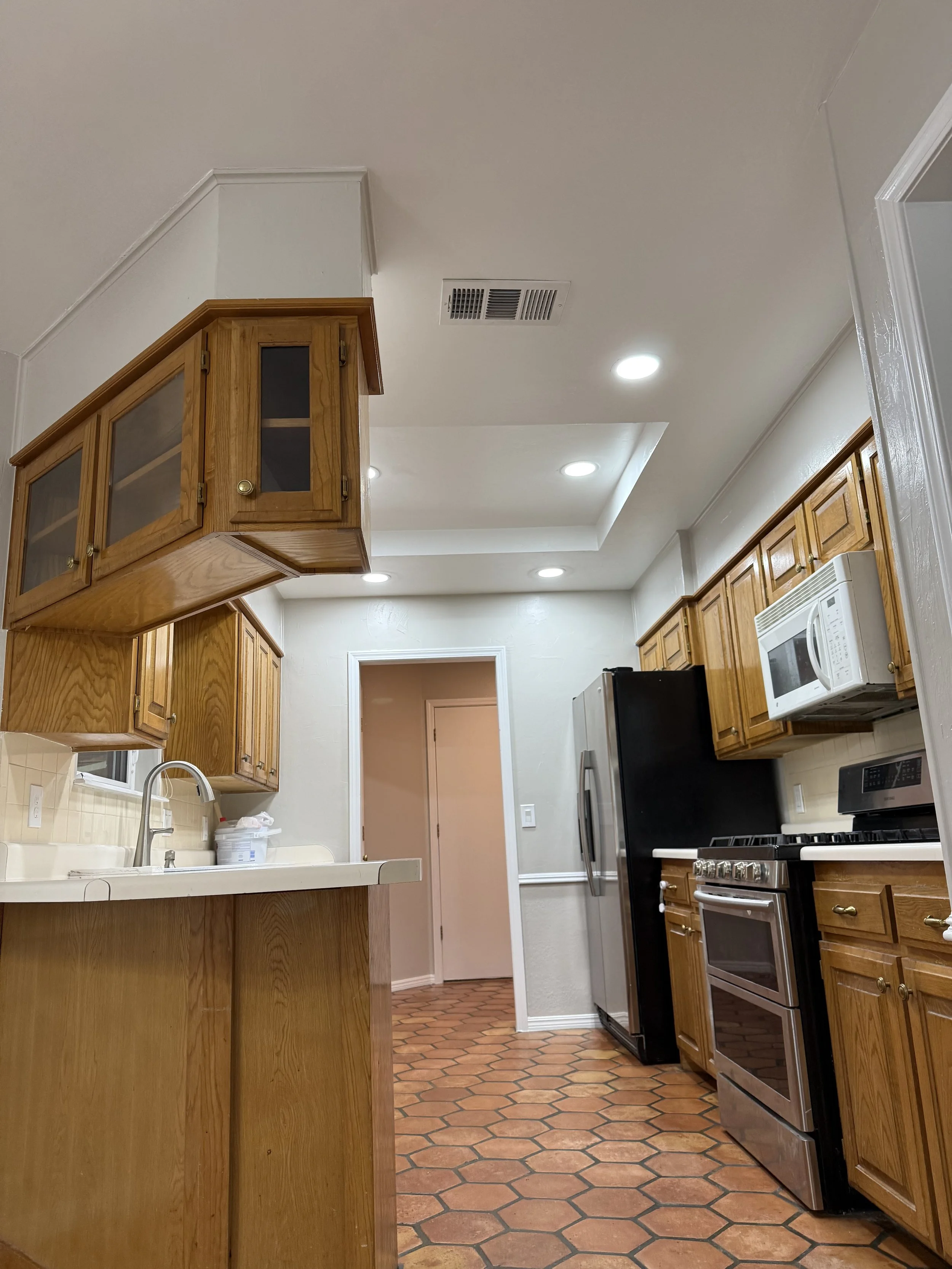 A kitchen with wooden cabinets, a black refrigerator, a white microwave, a gas stove, and a white sink. The floor has hexagonal terracotta tiles. There is a doorway leading to another room, and the ceiling has recessed lighting.
