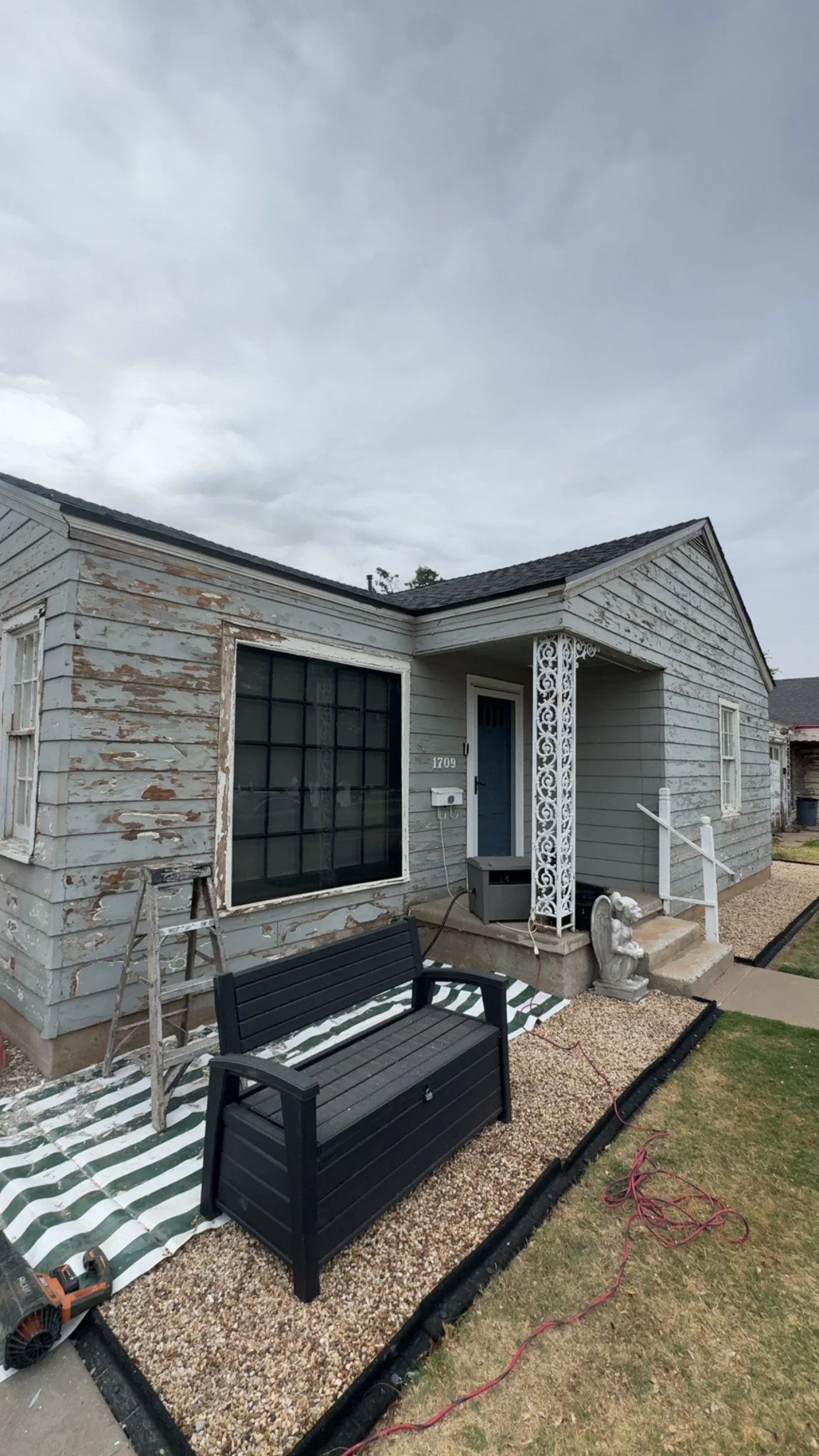 Front yard of a house with peeling gray paint, a large window, a blue door, and a decorative metal porch column. There are lawn furniture, gravel, a striped mat, and some tools and cords on the ground.
