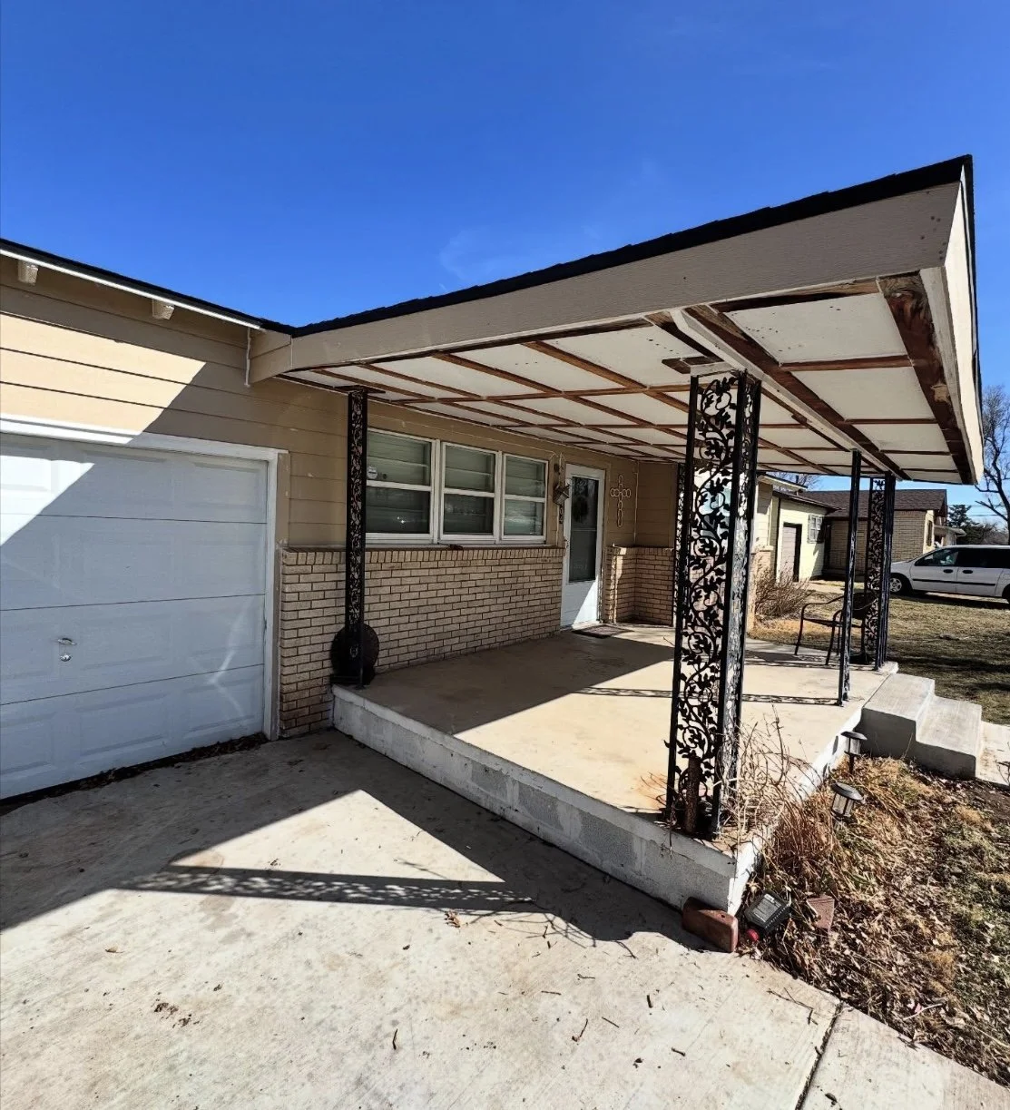 Front porch of a house with a concrete driveway, black decorative metal support columns, and a partially enclosed roof. The house has a brick and beige siding exterior with three small windows and a white garage door.