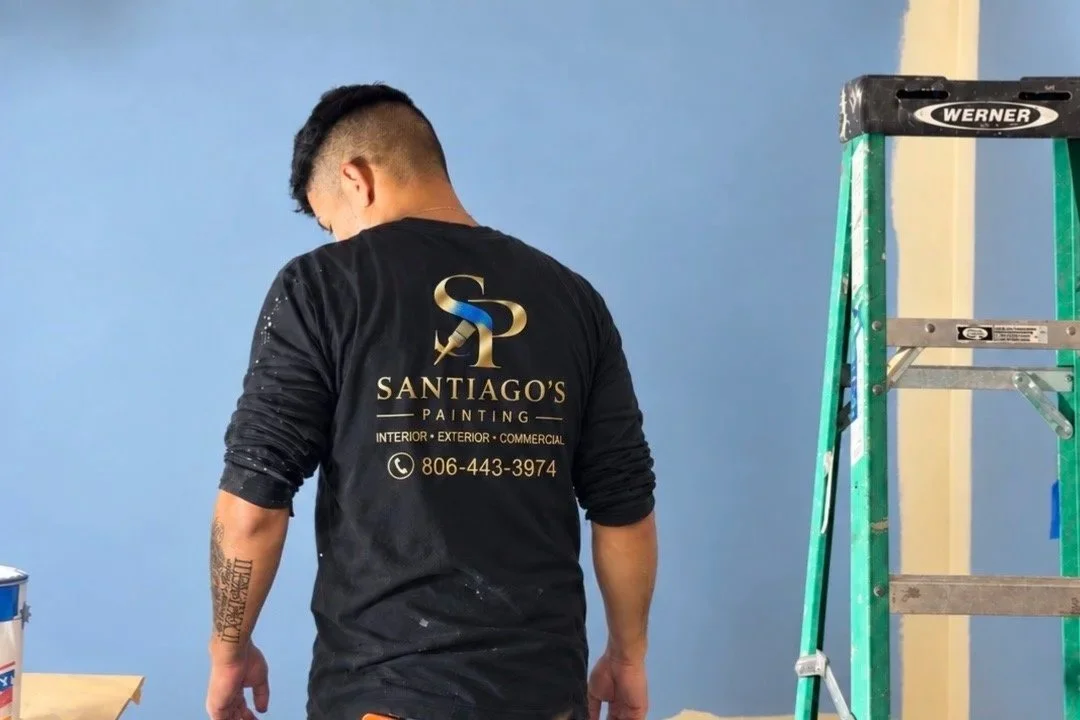 A man in a black shirt with gold lettering painting a wall blue, with a green ladder nearby.