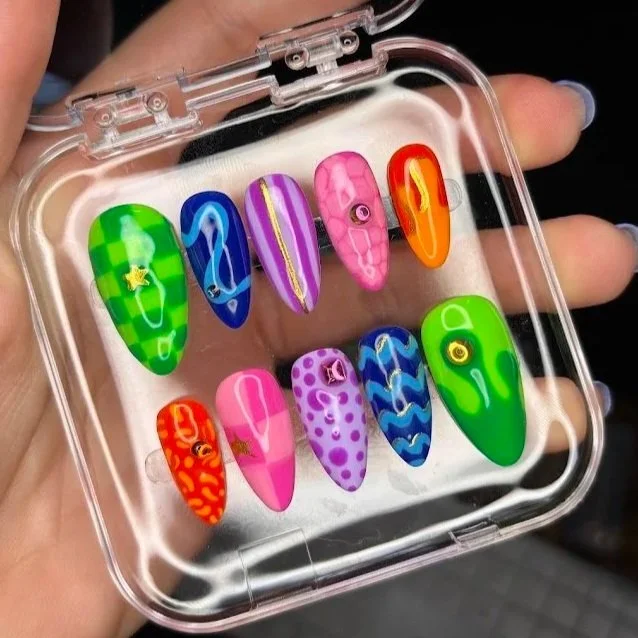 A hand holding a clear plastic box with a white foam insert, displaying ten colorful, decorated false nails inside.
