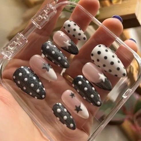 A hand holding a clear plastic case with ten oval-shaped decorated false nails, featuring black, white, and pink designs with stars and polka dots.