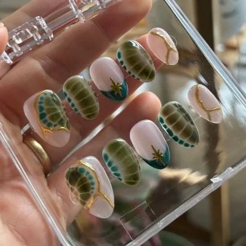 Hand holding a plastic case with decorative artificial nails featuring green gecko patterns and gold accents.