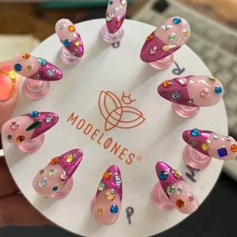 Display of colorful 3D nail art samples with various gemstones and decorations on pink plastic stands, arranged in a circle around a white surface with a 'Modelones' logo.