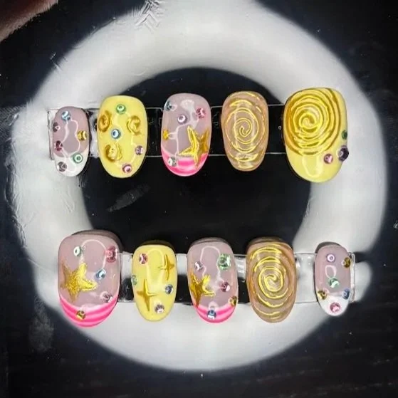 Collection of colorful decorated nail art nails arranged in two rows, featuring yellow, pink, and swirl patterns with small rhinestones and star designs.