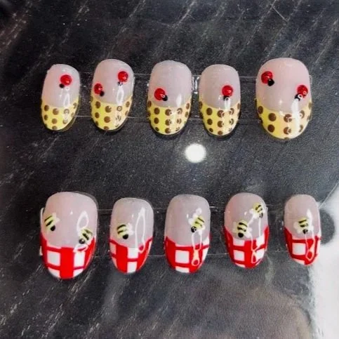 Nail art featuring a ladybug and bee theme, with some nails painted with red and yellow ladybug designs, and others with white background and bee illustrations, on a dark surface.