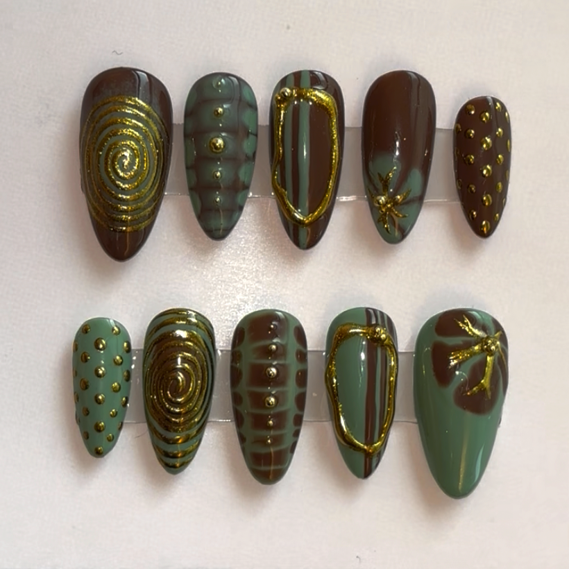 Set of ten artificial nail tips with green, brown, and gold design motifs, including spirals, dots, stripes, and dragonfly accents.