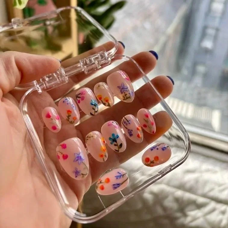 Hand holding a clear plastic case with decorative pressed flower nail decals inside near a window.