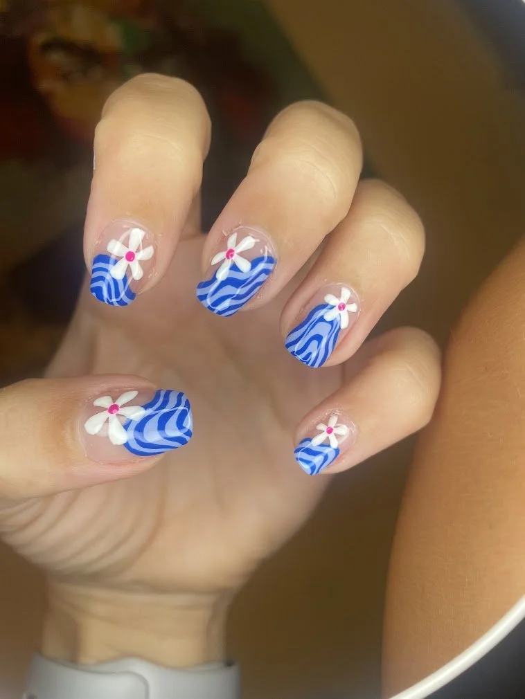 Close-up of a hand with painted nails featuring blue and white wavy patterns and white flowers with pink centers