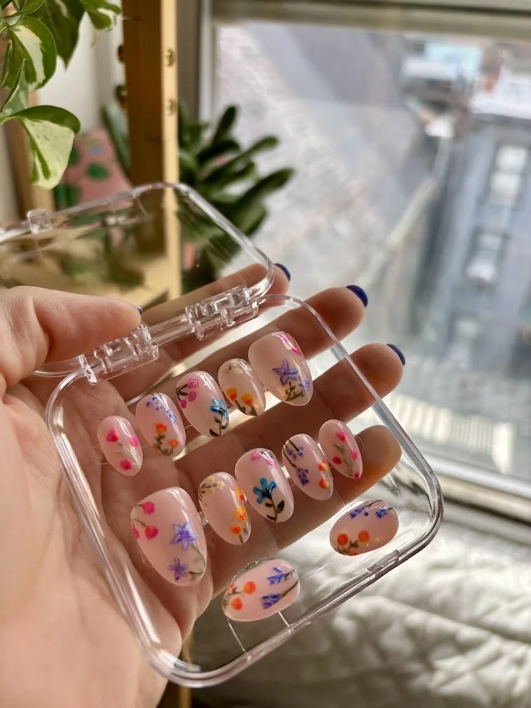 Hand holding a clear case with decorated false nails featuring floral and dot patterns near a window with an urban view.