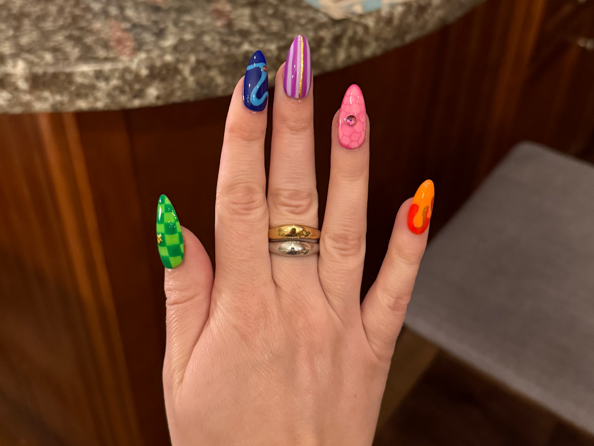 Hand with colorful, patterned acrylic nails, wearing two rings, against a wooden and granite background.