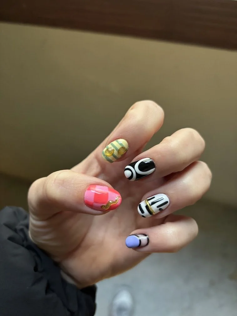Hand showing nails with colorful, decorative nail art designs including black and white patterns, pink and gold, yellow-green with gold, and blue with black accents.