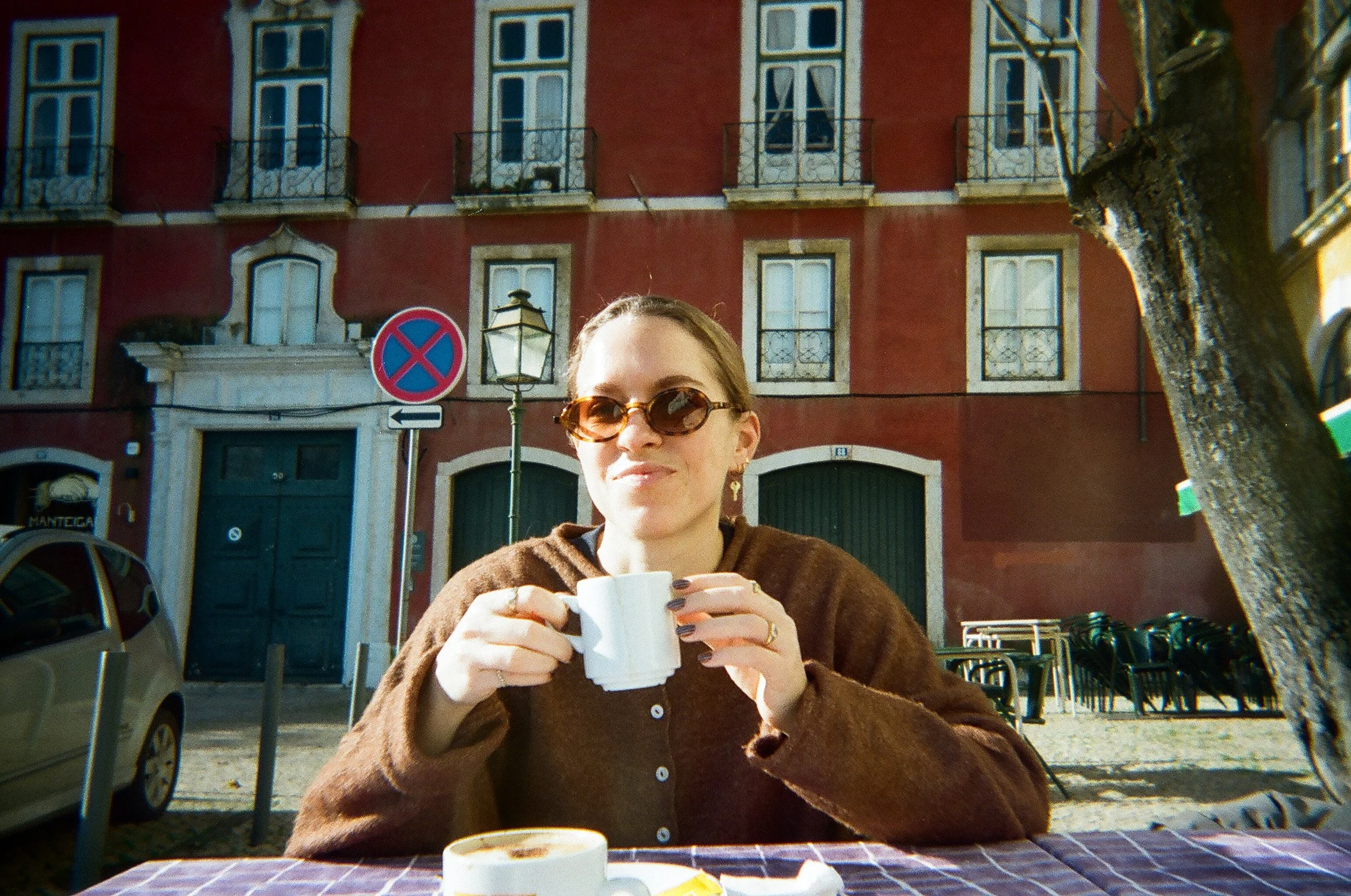 A woman with brown hair and sunglasses enjoying coffee at an outdoor cafe in front of a red building with multiple windows and balconies, a traffic sign, and a tree.