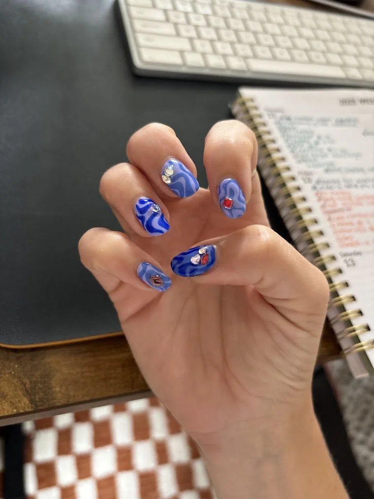 Close-up of a hand with blue marbled nail art featuring small rhinestone and gem accents, positioned over a desk with a keyboard, notebook, and tiled floor visible in the background.