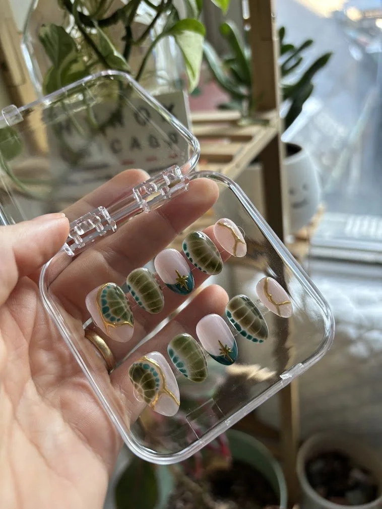 Hand holding a clear plastic container with decorative nail stickers featuring turtle shells and seashell designs, with indoor plants and a window in the background.