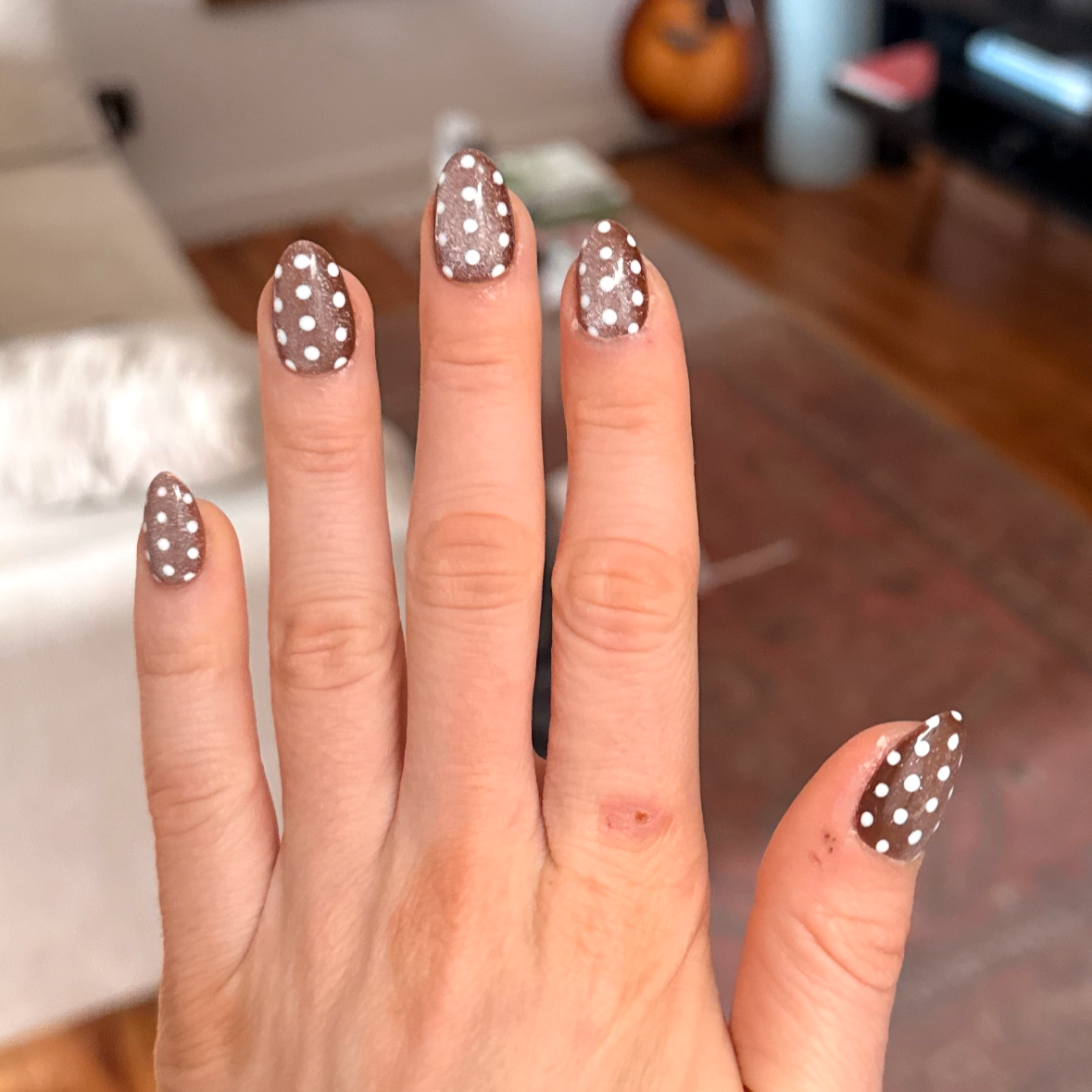 Close-up of a hand with manicured nails painted light brown with white polka dots, in a cozy room with wooden floors, a white couch, and a television in the background.