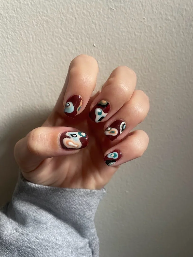 Hand with manicured nails painted in abstract swirl patterns with multiple colors against a textured beige wall.