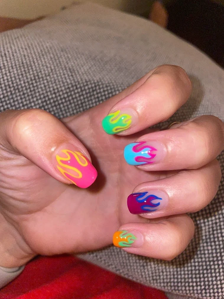 Close-up of a hand with colorful flaming nail art on each fingernail, featuring yellow, pink, blue, green, orange, and purple flames.