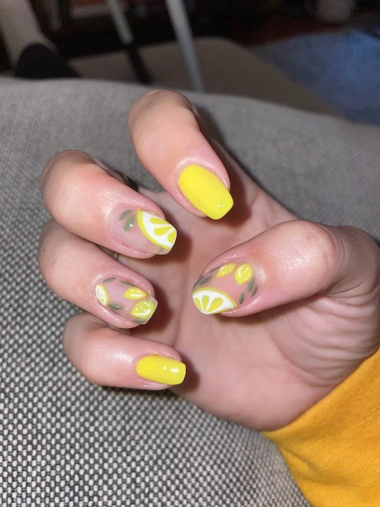 Manicured fingernails with yellow and white lemon slice nail art and yellow nail polish on some fingers, resting on a gray fabric surface.