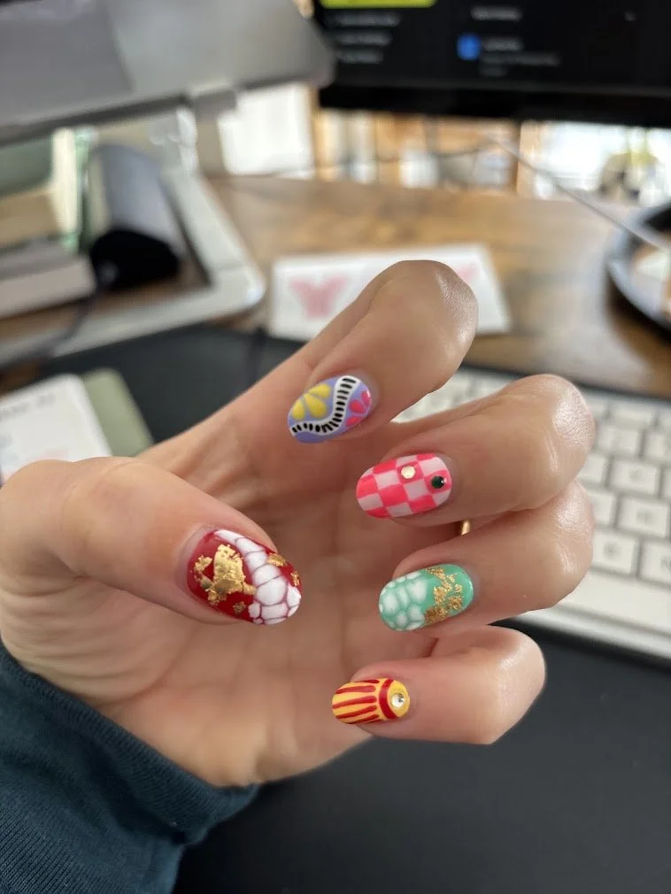 Close-up of a hand with colorful, intricate nail art featuring floral, checkered, and gold designs, held in front of a blurred workspace with a keyboard, computer screen, and office supplies.