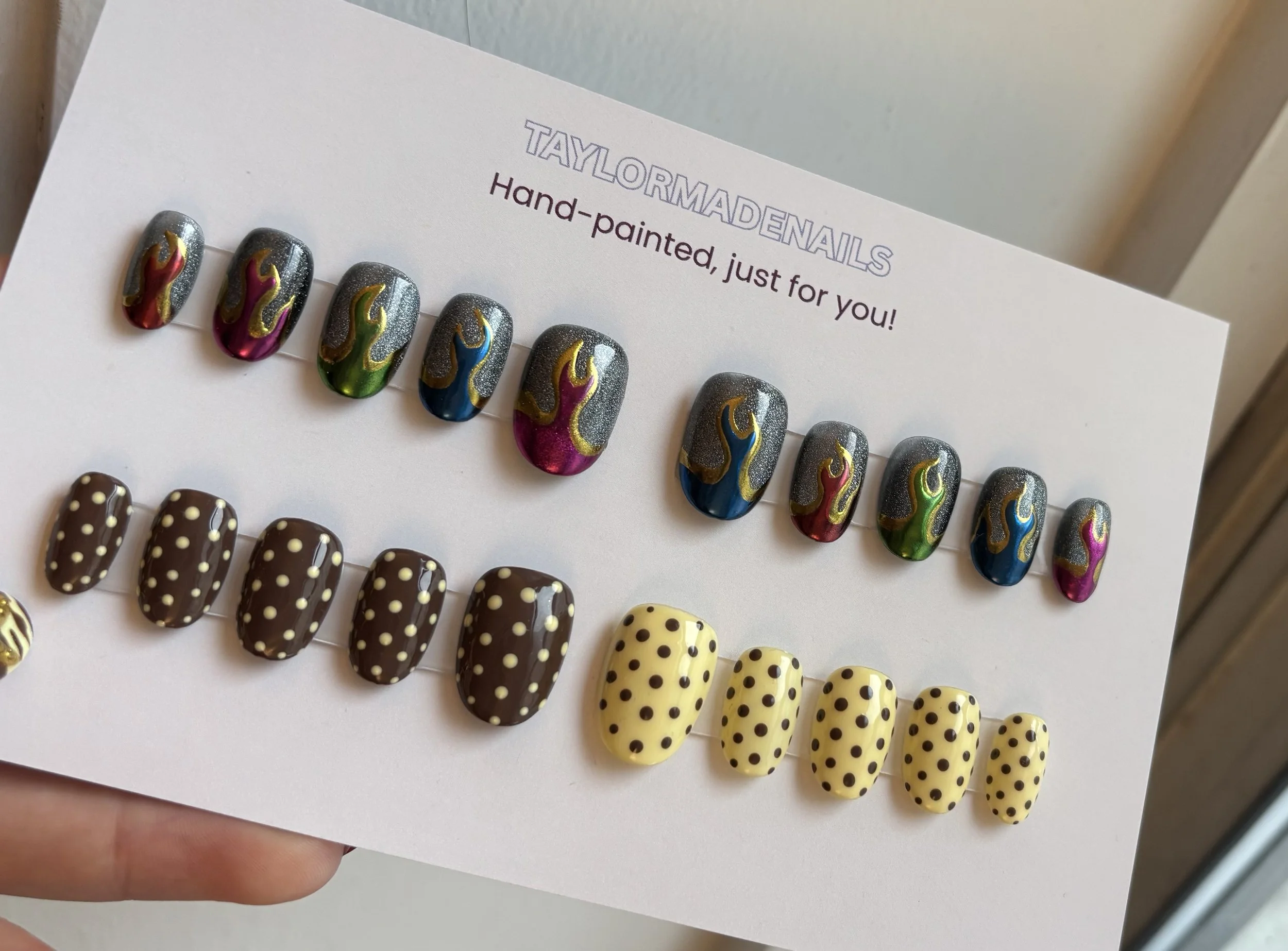 Close-up of a display card with colorful, flame-shaped nail art designs on artificial nails, arranged in a row. The card reads 'TAYLORMADENAILS' at the top, with a line of smaller text below that says, 'Hand-painted, just for you!' The background is 