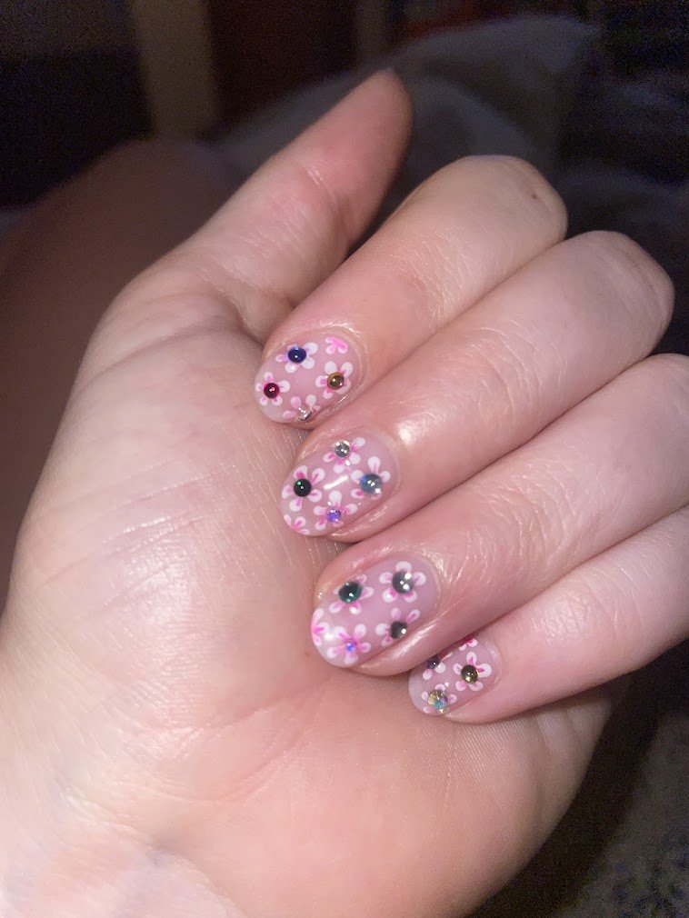 Close-up of a hand with decorated fingernails featuring pink, purple, black, green, and blue stones and white flower designs on a transparent pink base.