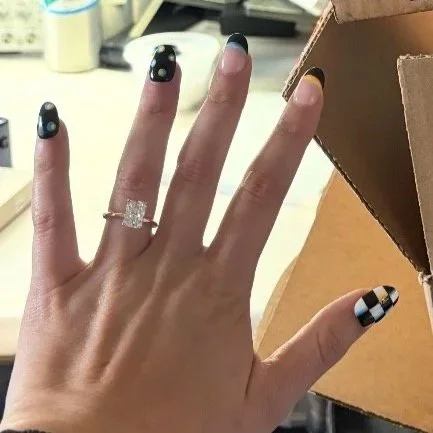 Close-up of a woman's hand with black and white checkered nail polish, wearing a large square diamond ring on her ring finger, holding a cardboard box in an indoor setting.
