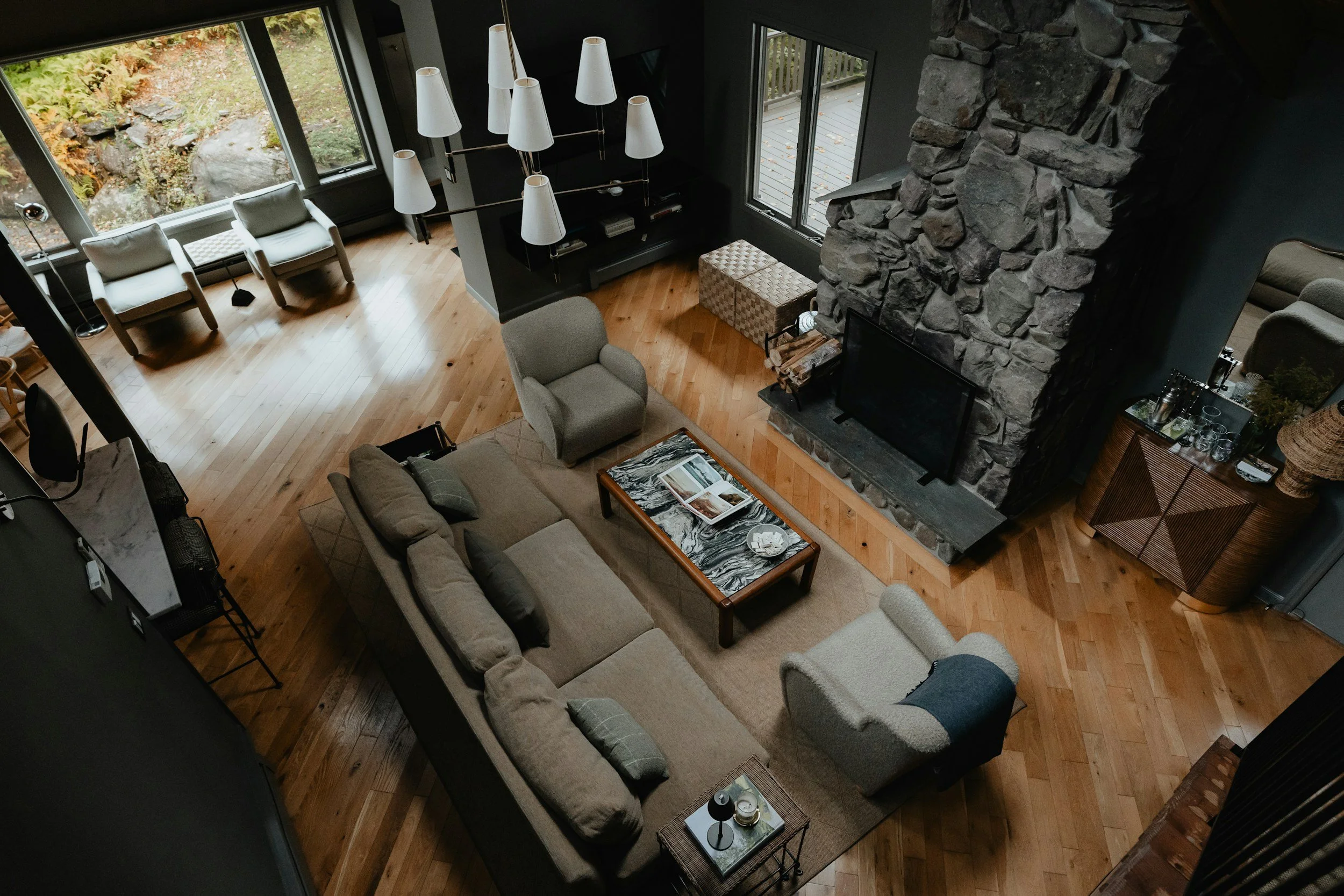 A spacious living room with wooden floors, large windows showing outdoor greenery, a stone fireplace, beige sofas, a gray armchair, a patterned ottoman, and a chandelier with multiple white shades.