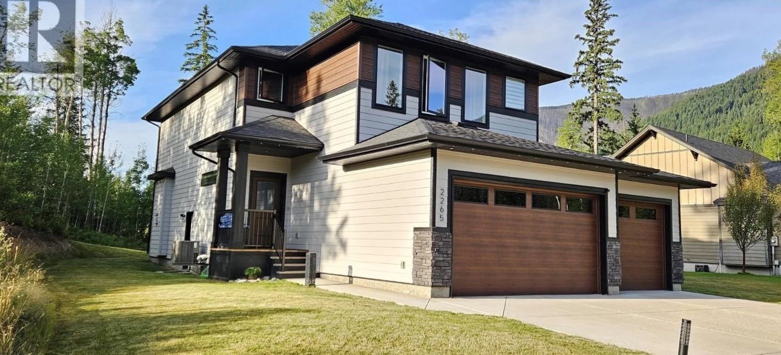 Modern two-story house with white siding and dark brown accents, featuring a two-car garage and surrounded by green lawn and trees.